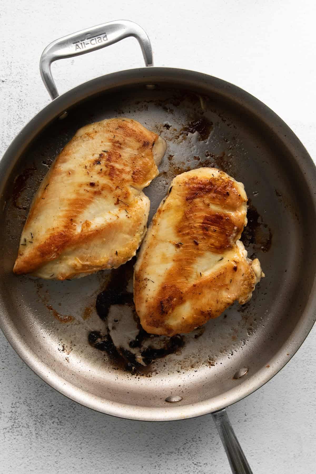 Two cooked chicken breasts in a stainless steel skillet. The chicken has a golden-brown sear and the pan shows some residual cooking marks.