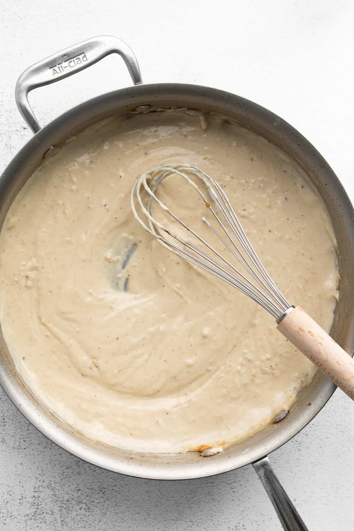 A metal whisk rests in a large, stainless steel pan filled with creamy, beige sauce.