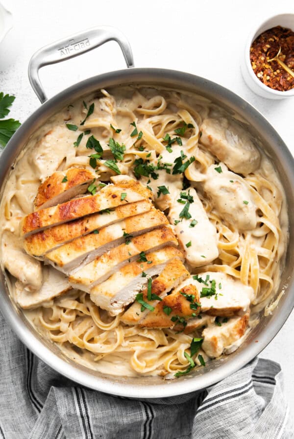 A pan filled with creamy fettuccine pasta topped with slices of grilled chicken and garnished with parsley. A small bowl of red pepper flakes is seen in the background.