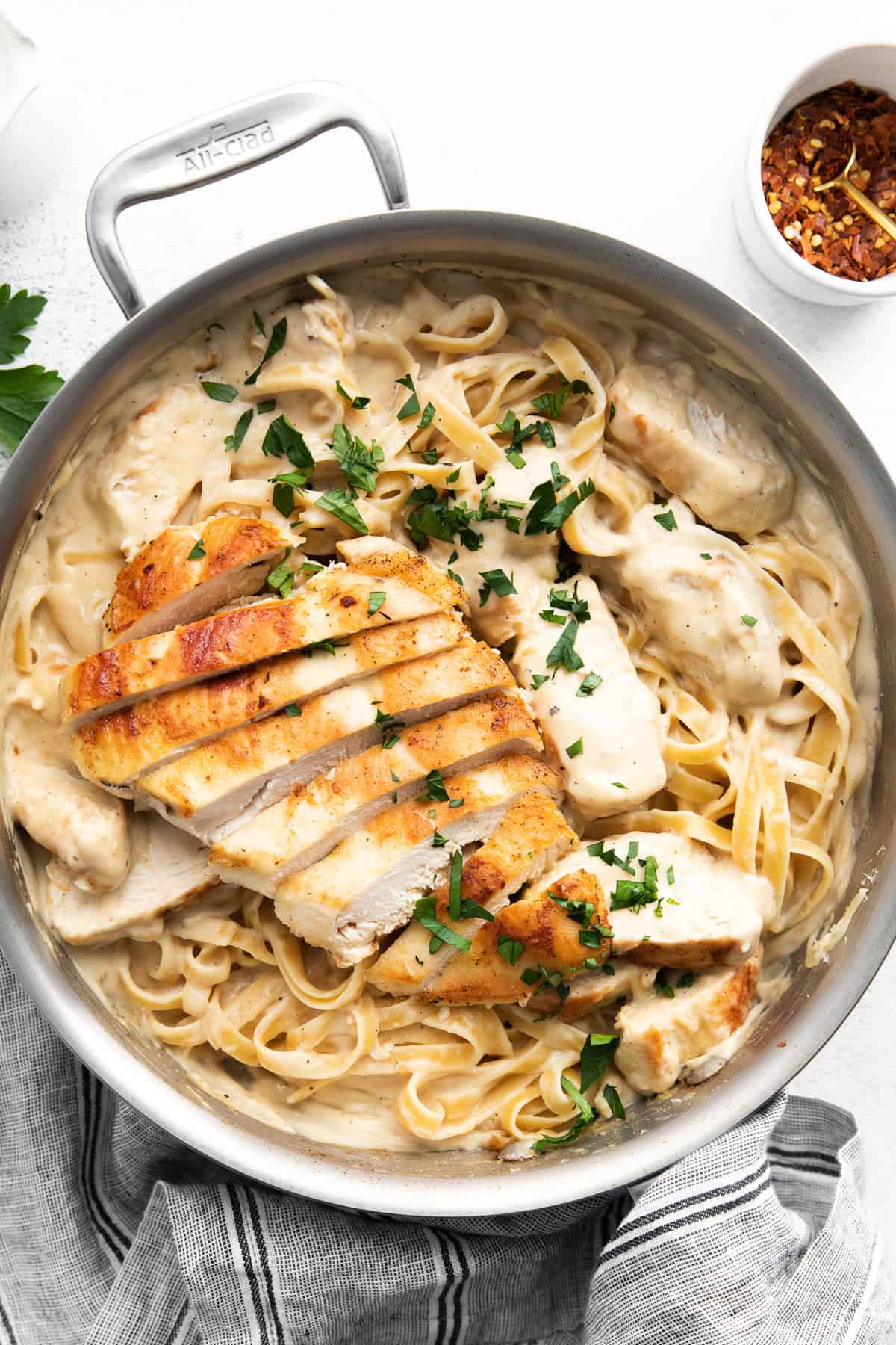 A pan filled with creamy fettuccine pasta topped with slices of grilled chicken and garnished with parsley. A small bowl of red pepper flakes is seen in the background.