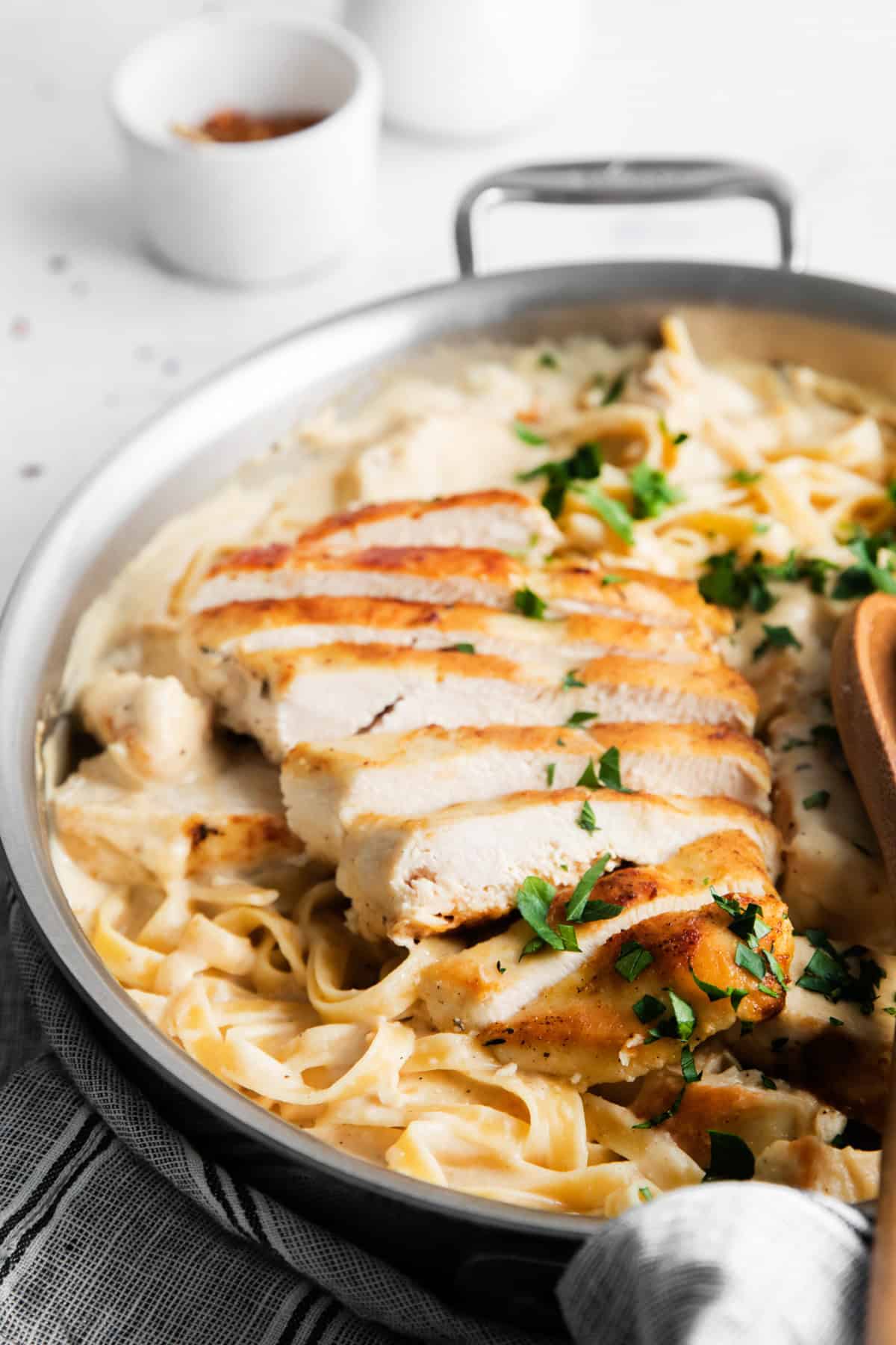 A skillet containing creamy fettuccine Alfredo topped with sliced grilled chicken and garnished with chopped parsley.