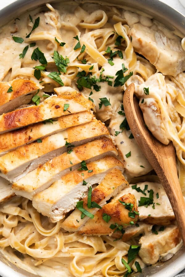 A skillet filled with creamy fettuccine Alfredo topped with sliced grilled chicken and garnished with chopped parsley. Wooden utensils are placed on the side.