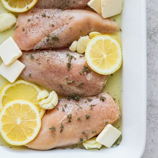 chicken breasts with lemon slices and herbs in a white baking dish.