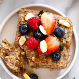 A square piece of baked oatmeal topped with sliced almonds, strawberries, raspberries, and blueberries sits invitingly on a white plate beside a fork.