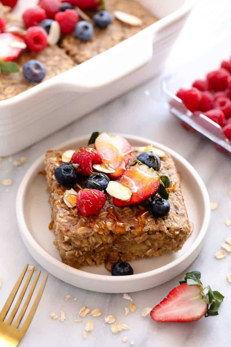 A slice of baked oatmeal, adorned with strawberries, raspberries, blueberries, and almond slices, rests on a white plate. Nearby, a gold fork glistens beside scattered oats and a dish of vibrant berries.