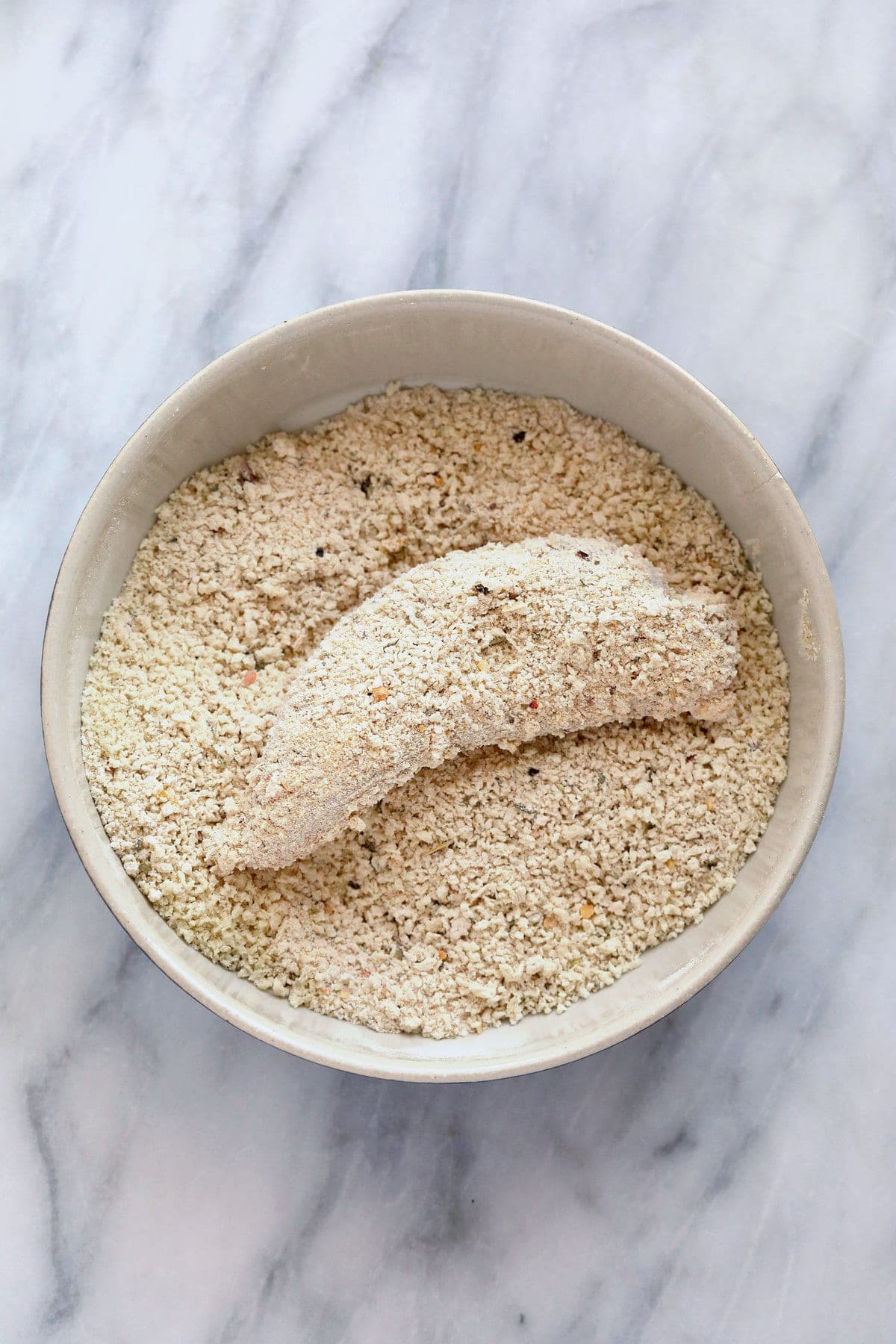 A chicken tender in a bowl of bread crumbs and spices. 