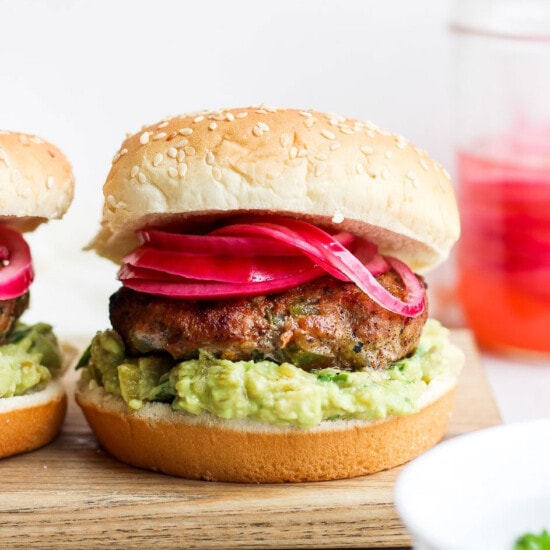 two burgers with guacamole and red onion on a cutting board.