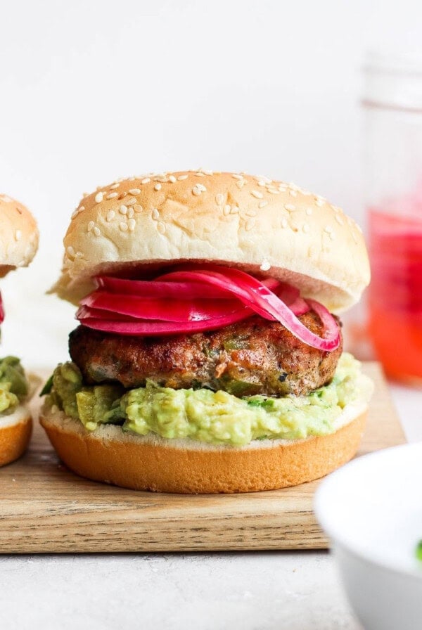 two burgers with guacamole and red onion on a cutting board.