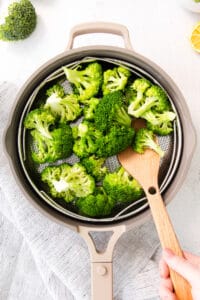 a person is slicing broccoli in a pan with a wooden spoon.