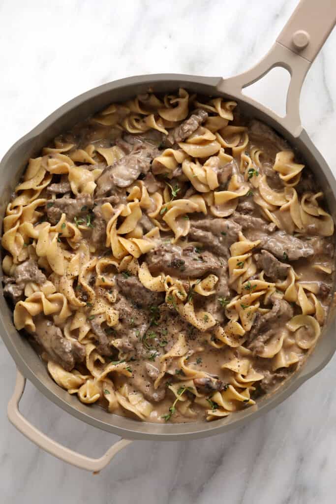 Classic beef stroganoff in a pan with fresh thyme, ground pepper, and egg noodles. 