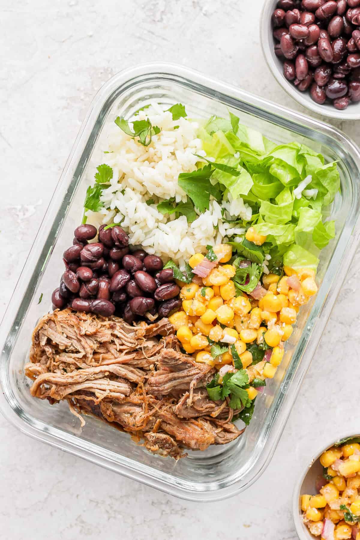 A slow cooker carnitas meal with rice, beans, and corn served in a glass container.