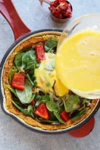 Pouring egg mixture into a skillet with spinach, red bell peppers, and sweet potato crust for quiche preparation.