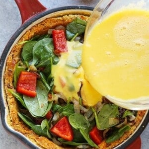Pouring egg mixture into a skillet with spinach, red bell peppers, and sweet potato crust for quiche preparation.