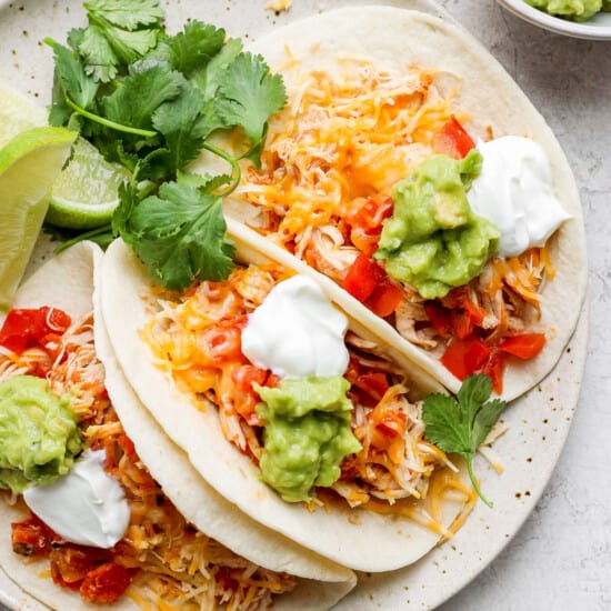 three chicken tacos on a plate with guacamole and sour cream.