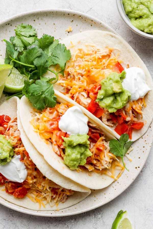 three chicken tacos on a plate with guacamole and sour cream.