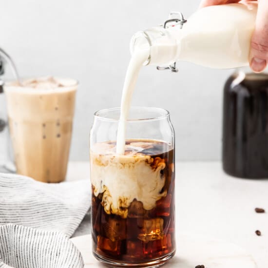 a person pouring milk into a glass of iced coffee.