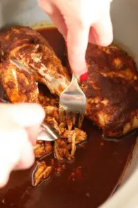 A person prepares slow cooker pulled chicken by pouring sauce over it.
