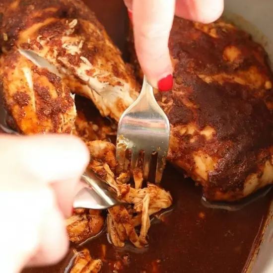 A person prepares slow cooker pulled chicken by pouring sauce over it.