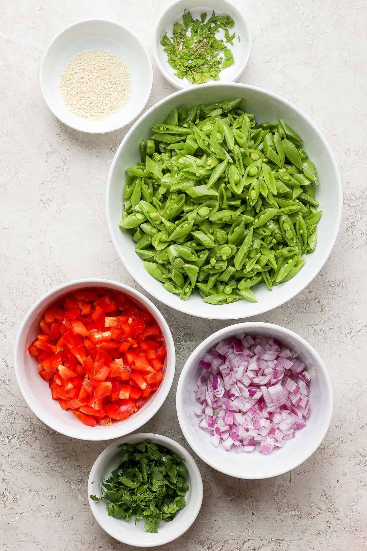 Sugar snap peas, red peppers, red onion, sesame seeds, and fresh mint in small bowls. 