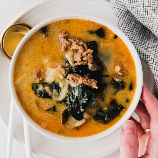 A person holding a bowl of zuppa toscana with meat and greens.