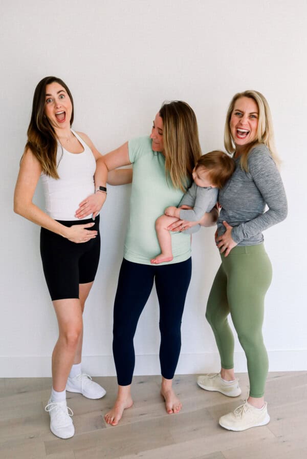 A group of women wearing lululemon maternity clothes are standing in front of a white wall with a baby in their arms.