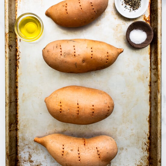 Four sweet potatoes on a baking sheet with olive oil and spices.