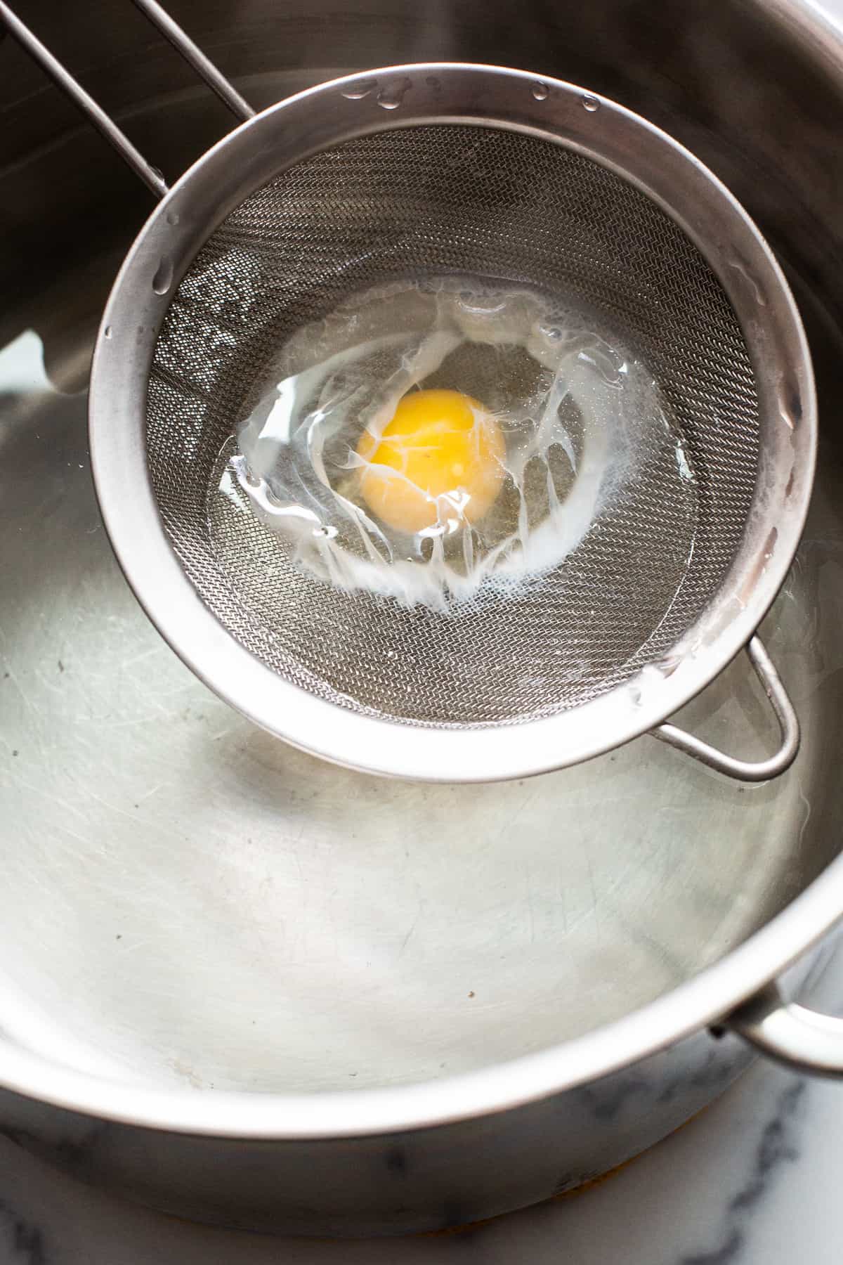 Raw egg in a mesh strainer being put in a pot of boiling water.