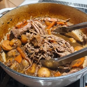 A pot of savory pot roast with tender shredded beef, carrots, and potatoes in flavorful broth. Tongs rest on the edge of the pot.