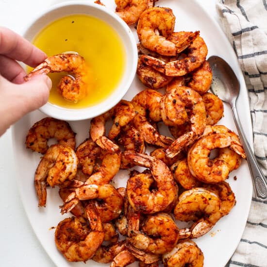 A plate of grilled shrimp with a hand holding a bowl of olive oil.
