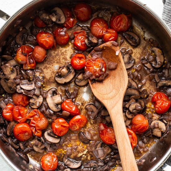 Mushrooms and tomatoes in a pan with a wooden spoon.
