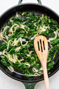 Spinach and onions in a skillet with a wooden spoon.