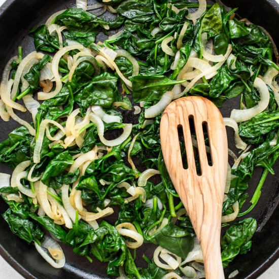 Spinach and onions in a skillet with a wooden spoon.