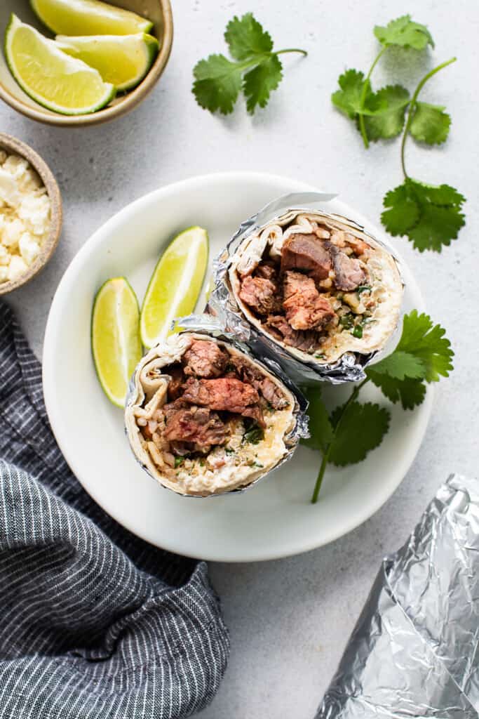 Steak burrito on a plate with sliced limes and cilantro.