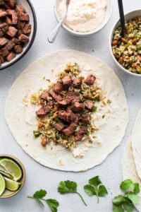 Beef tacos on a white table with limes and garnishes.