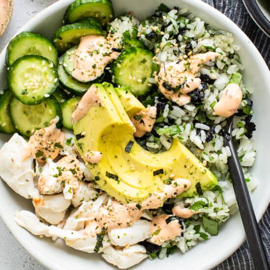 A white bowl with rice, cucumbers and avocado.