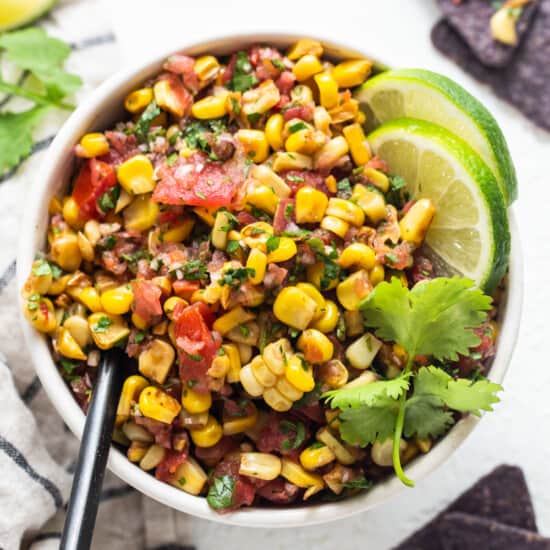 mexican corn salad in a white bowl with limes and tortilla chips.