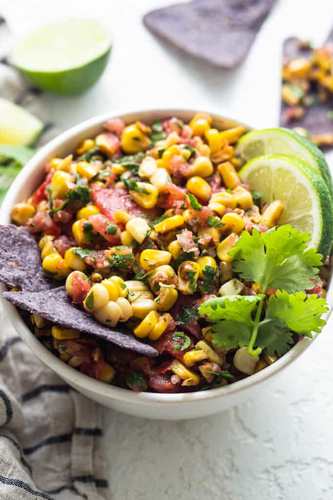 mexican corn salsa in a white bowl with limes and tortilla chips.