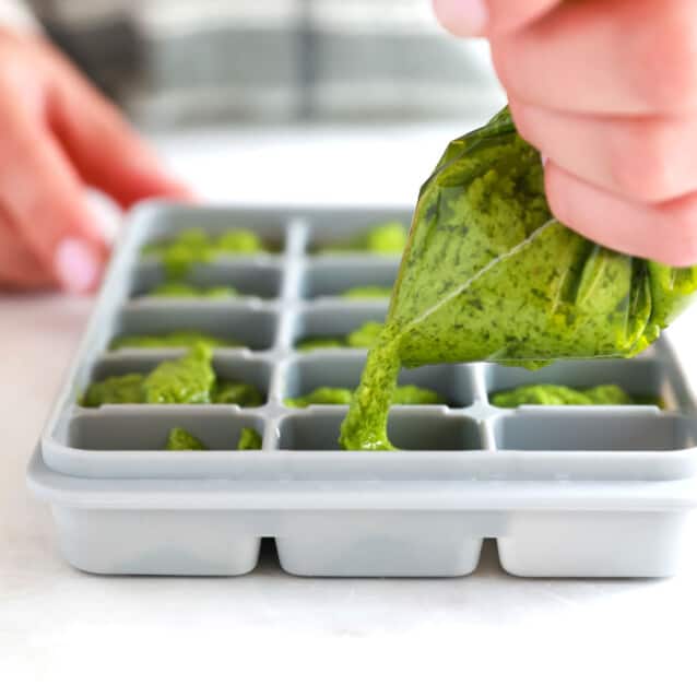 A person preparing homemade pesto by pouring it into an ice cube tray.