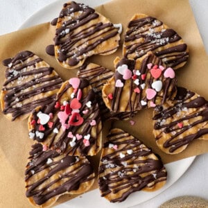 Heart-shaped cookies drizzled with chocolate, sprinkled with salt, and adorned with heart-shaped sprinkles rest on a parchment-lined plate. Nestled beside them are delectable peanut butter cups, adding an irresistible touch to this sweet display.