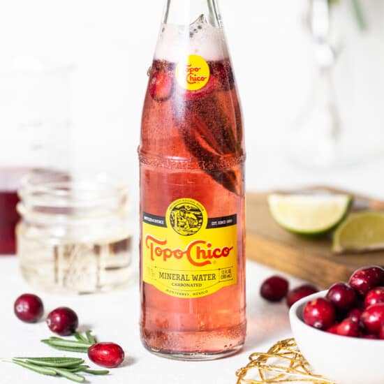 A bottle of cranberry soda with cranberries and sprigs of rosemary.