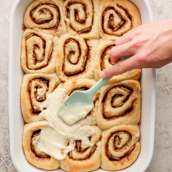 A person is dipping a spatula into a dish of cinnamon rolls.