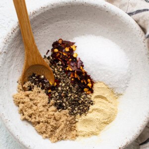 A bowl holds a savory steak seasoning mix of brown sugar, black pepper, crushed red pepper flakes, salt, and garlic powder, with a wooden spoon resting on the side.