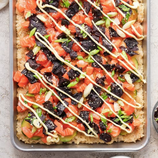 A pizza with salmon, avocado, and arugula on a baking sheet.