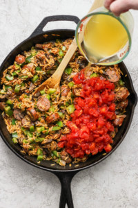 A person pouring oil into a skillet with rice and sausage.