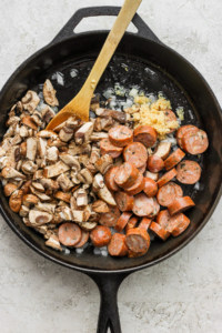 Sausages and mushrooms in a skillet with a wooden spoon.