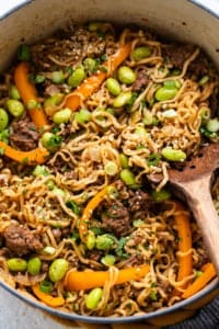 A pan full of noodles and vegetables with a wooden spoon.