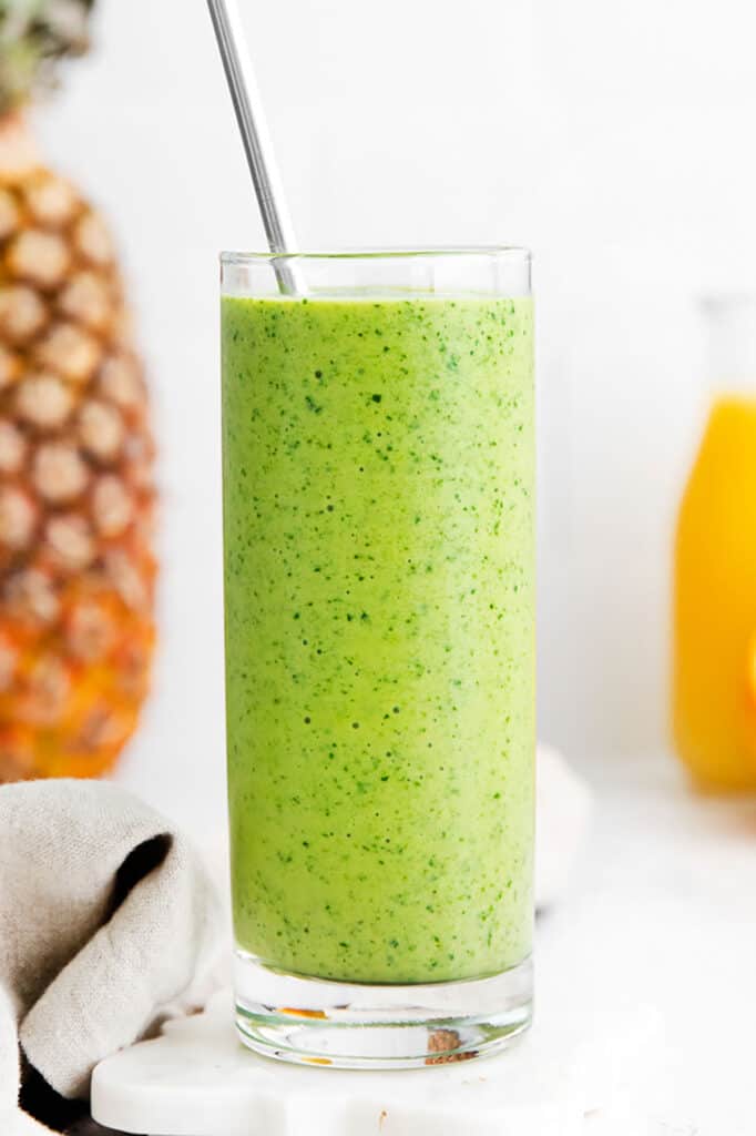 A spinach smoothie in a glass next to oranges and pineapples.