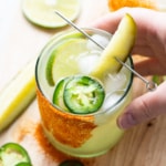 A hand holding a glass of drink with a pickle and lime.