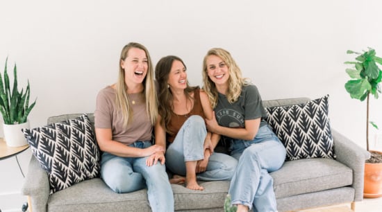 Three women laughing together on a sofa in a bright, cozy living room.