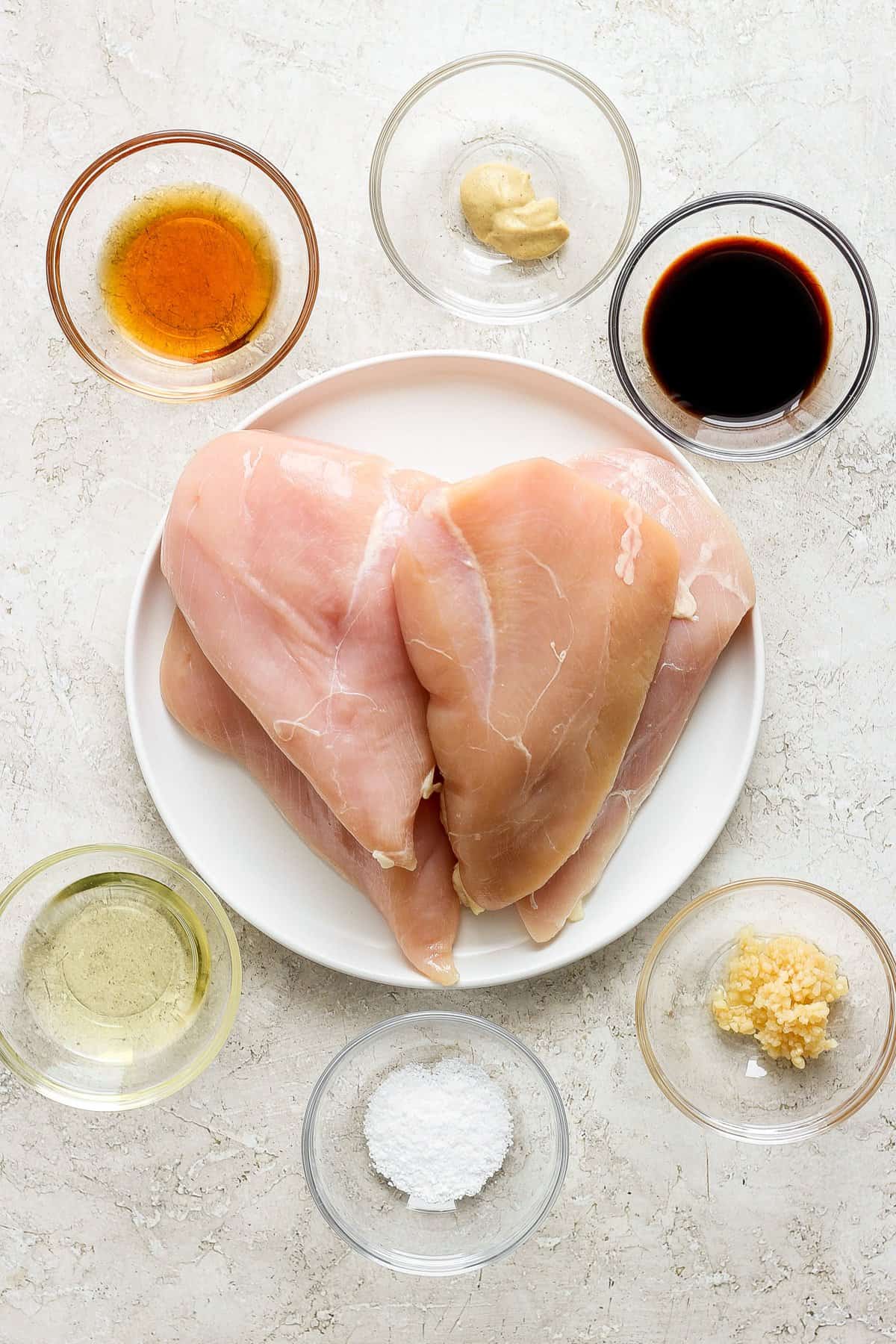 Raw chicken breasts on a plate with various seasonings and marinade ingredients arranged around it.